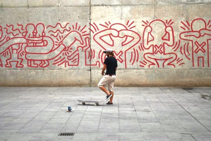 Man standing on his skateboard in front of a red Keith Haring mural on a concrete wall in Barcelona.