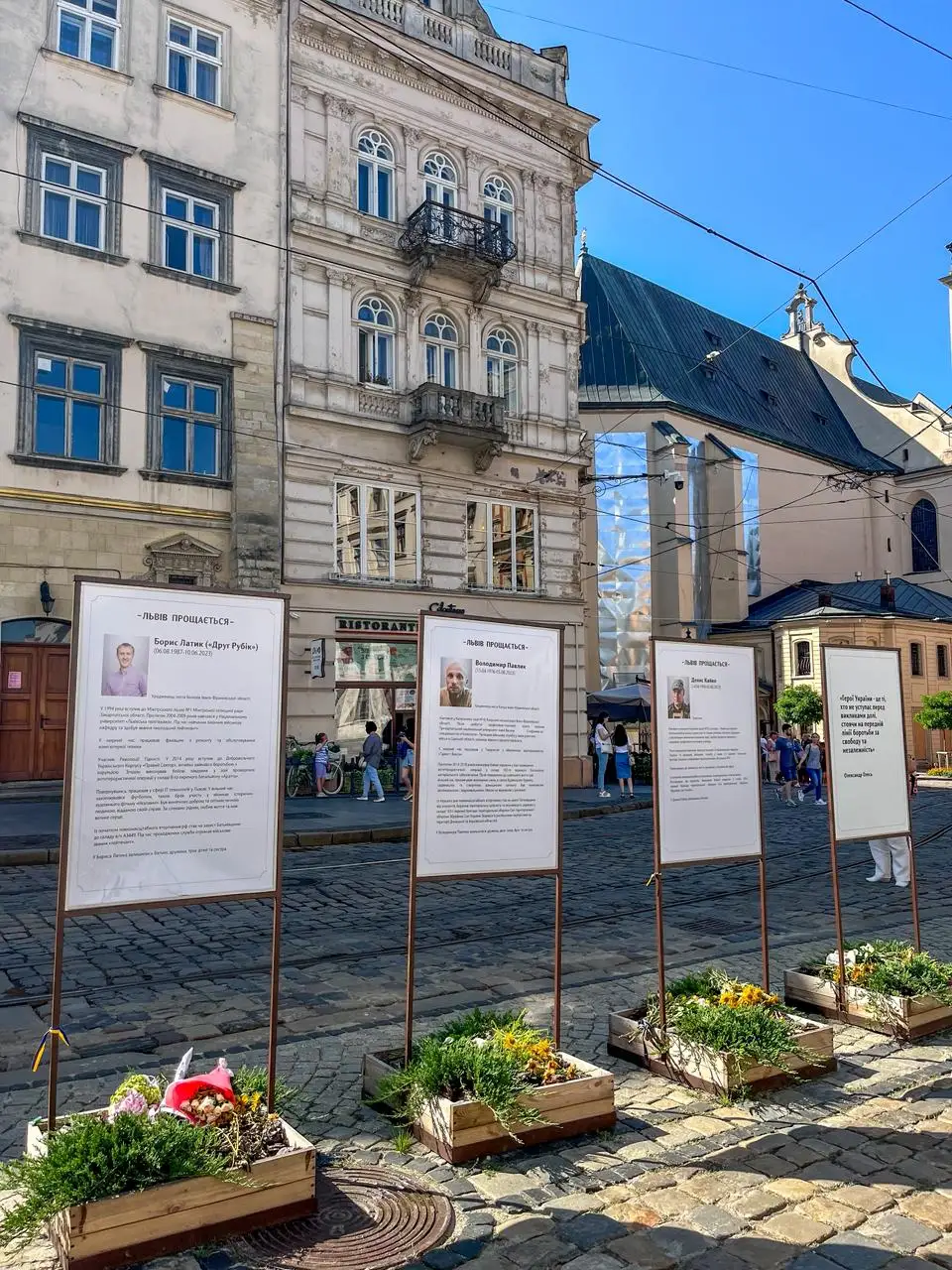Four memorial stands with portraits and text on a cobblestone street in Lviv, with historic buildings and a church in the background.