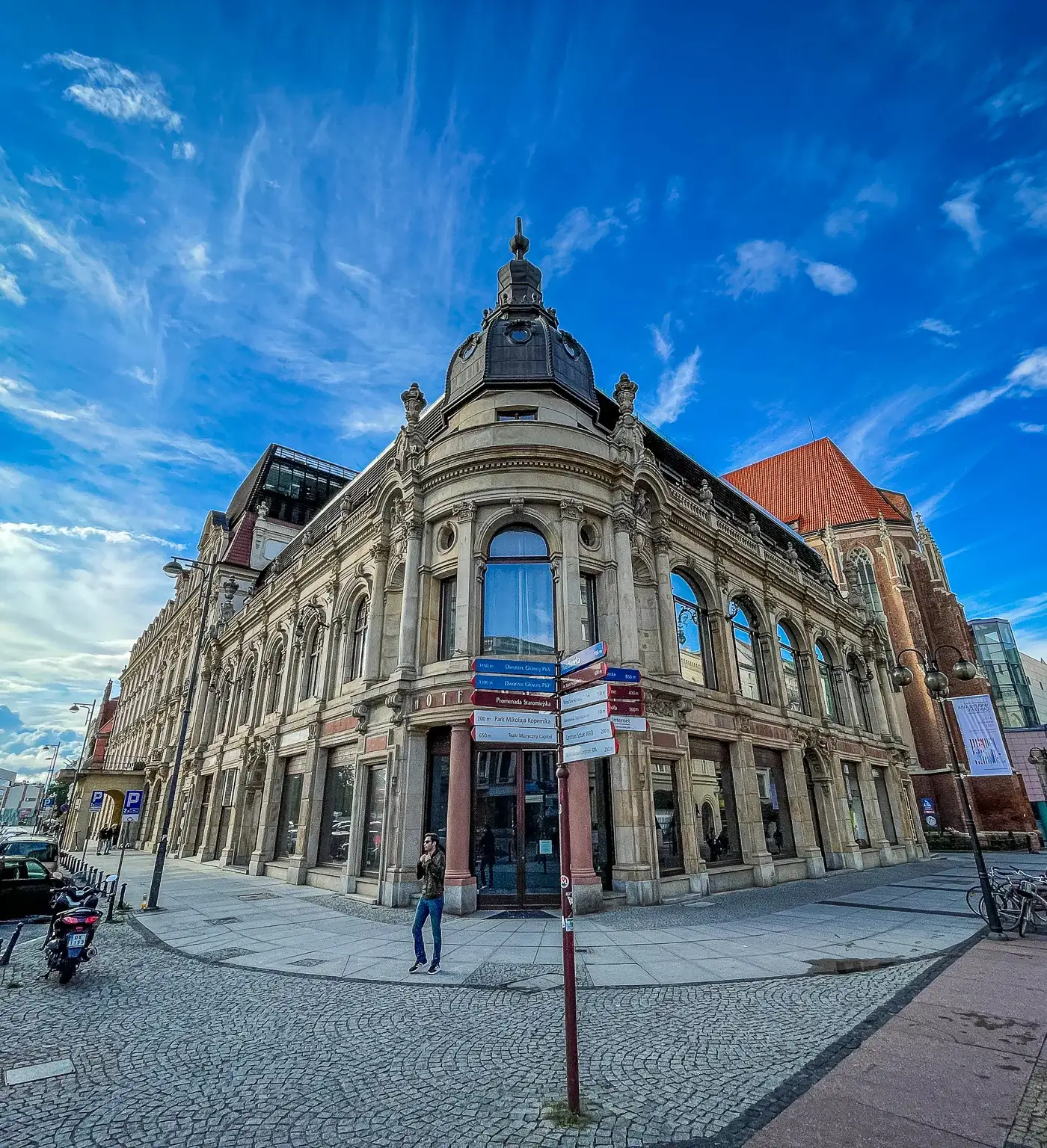 Ornate historic building with a domed corner tower and large windows on a cobblestone street in Wrocław. A street signpost is visible.