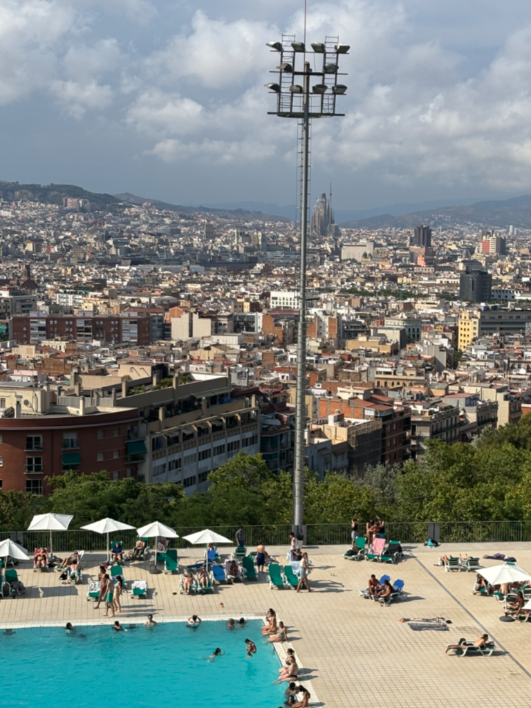 Panoramic view of Barcelona city, featuring a swimming pool and sunbathers in the foreground, with Sagrada Familia in dist...