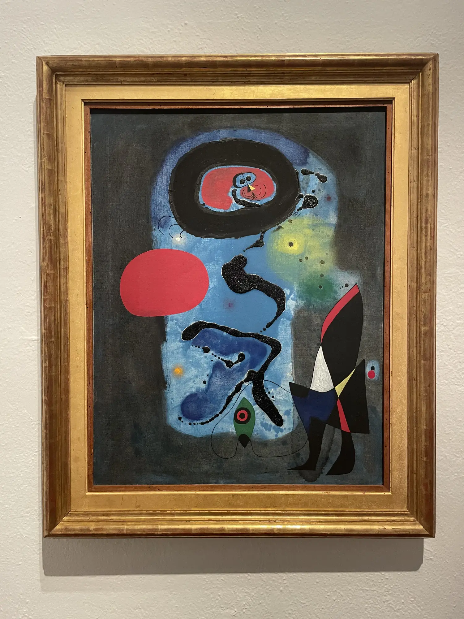 Abstract painting by Joan Miró in a gold frame, featuring primarily blue, black, and red shapes on a dark background.