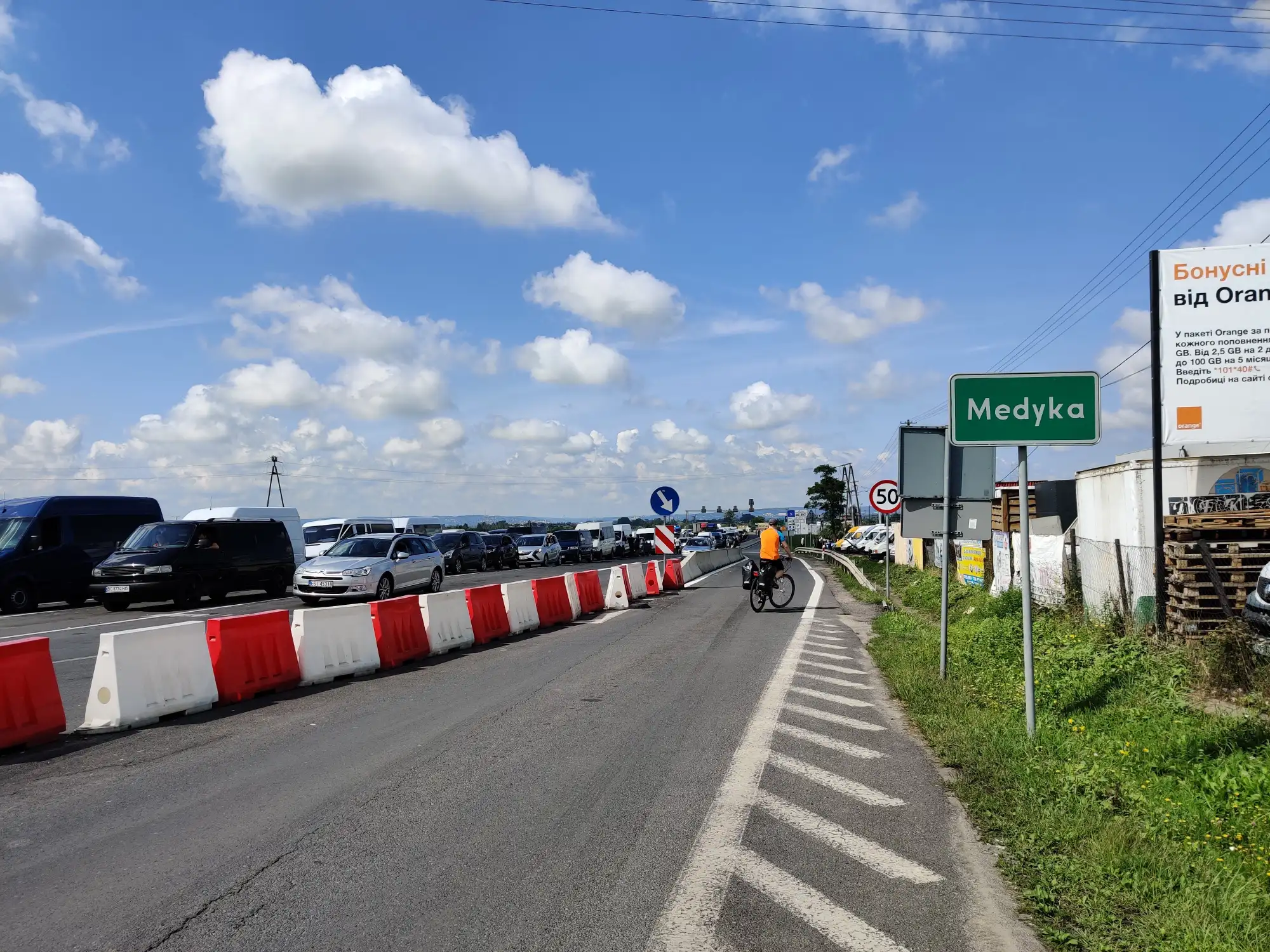 Droga do przejścia granicznego w Medyce, kolejka samochodów, rowerzysta i tablica z nazwą miejscowości pod błękitnym niebem.