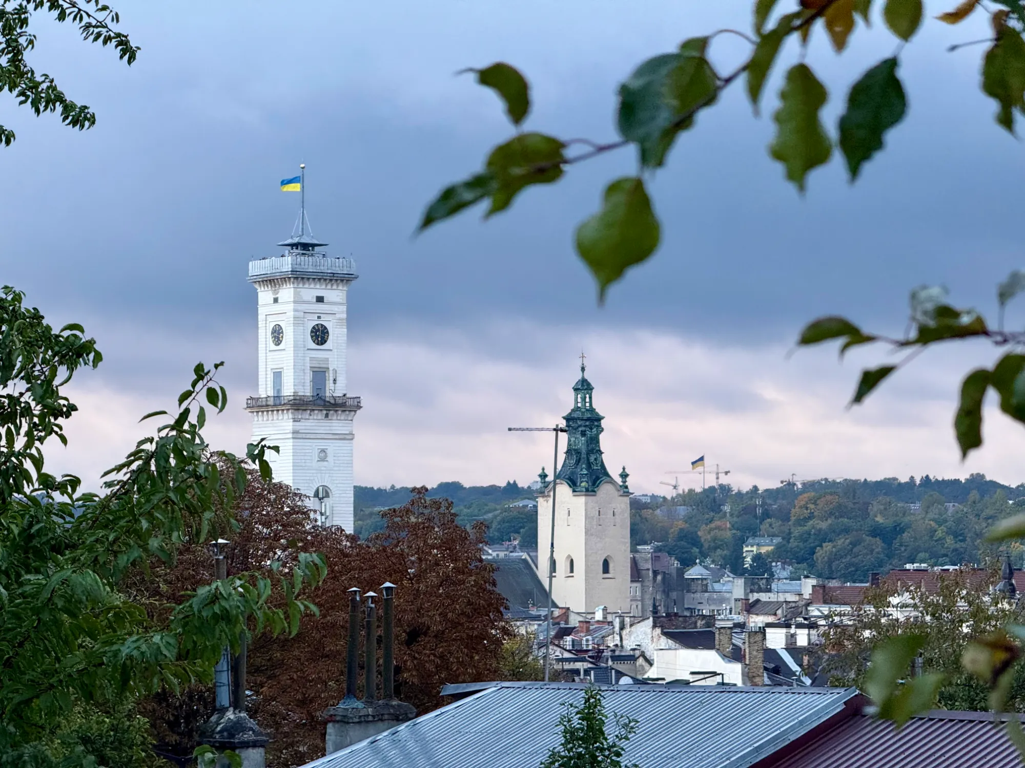 Widok na białą wieżę lwowskiego Ratusza i iglicę Katedry Łacińskiej nad dachami miasta pod zachmurzonym niebem.