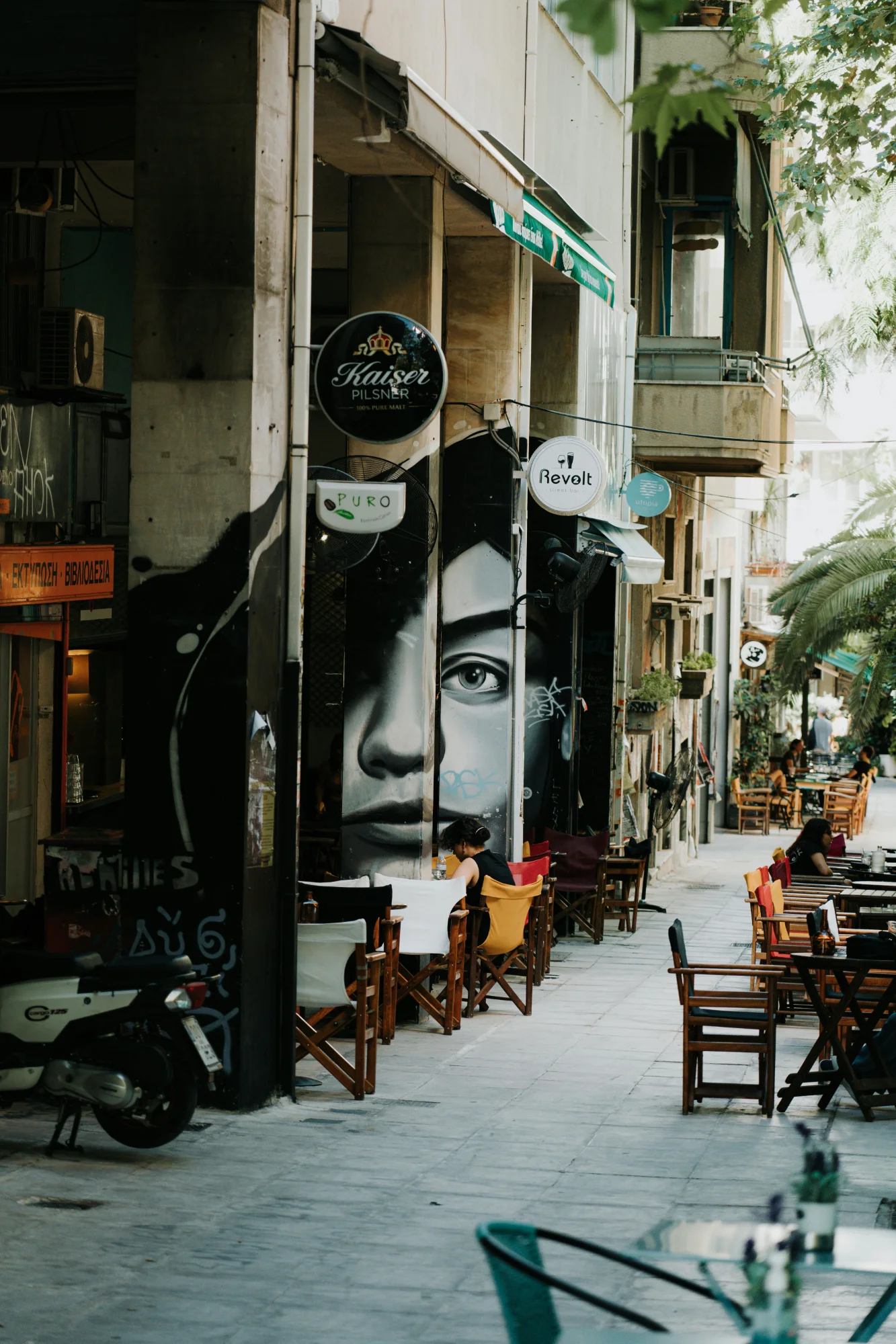 Street scene in Exarchia, Athens, with outdoor cafe seating, a scooter, and large monochrome face murals on pillars.