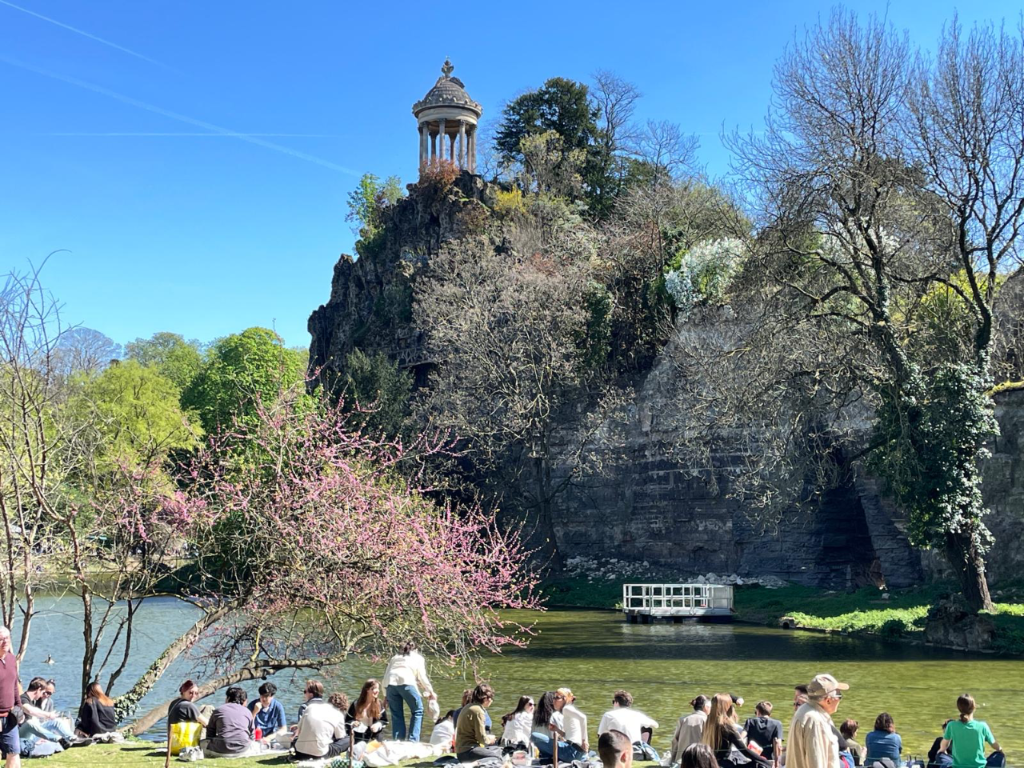 People relaxing on grass by a lake in Parc des Buttes-Chaumont, with the Temple de la Sibylle atop a rocky cliff in Paris.