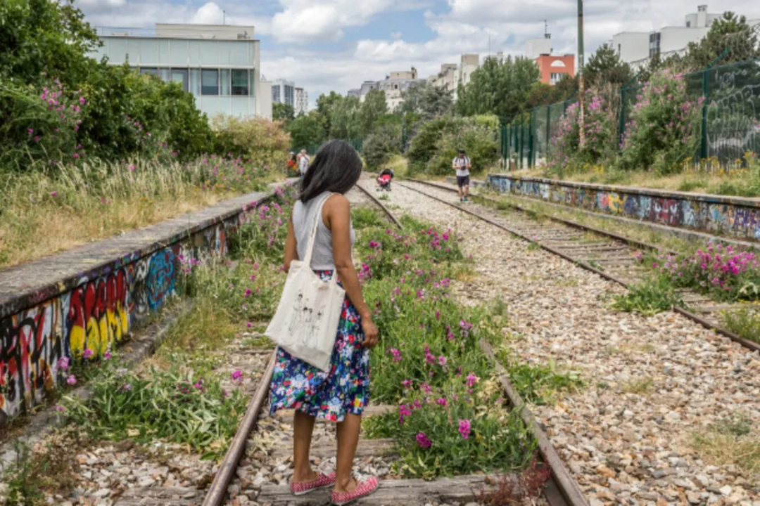 A woman walks along the abandoned Petite Ceinture railway tracks in Paris, surrounded by wildflowers and graffiti.