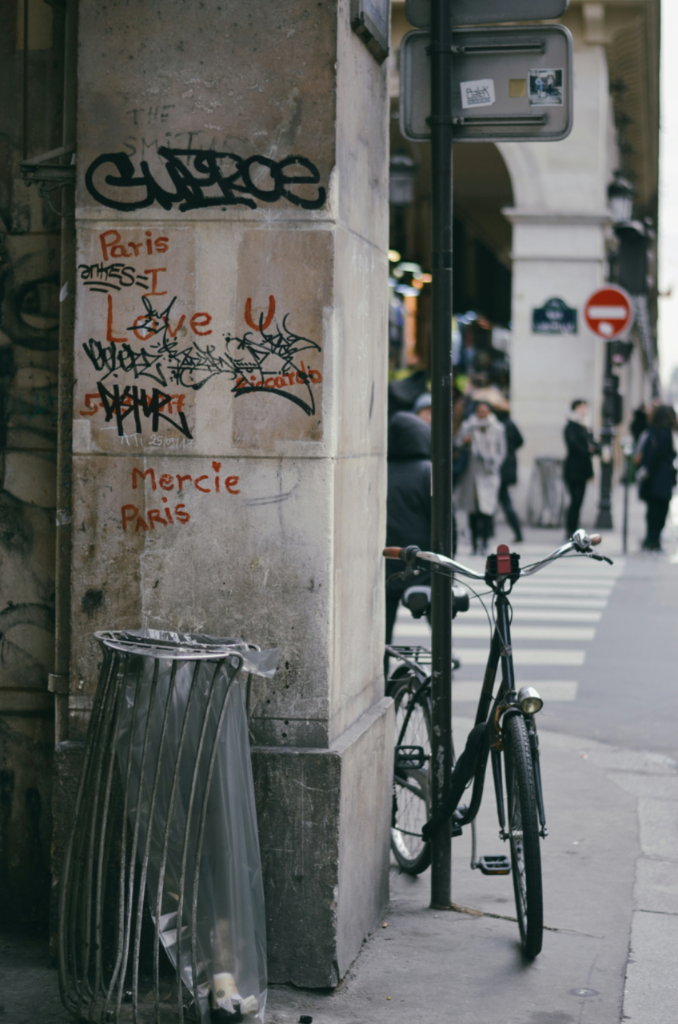 Stone pillar with Paris I Love U graffiti next to a wire trash bin and a parked bicycle on a city street.