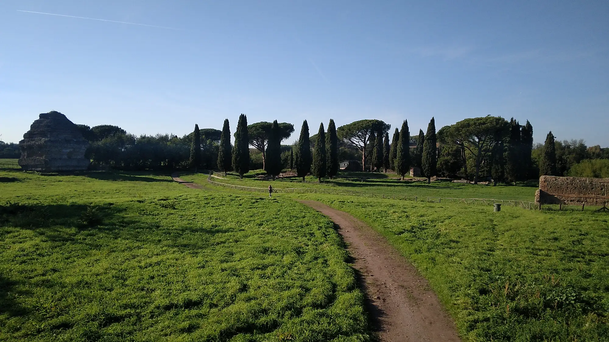 Path winding through Appian Way Park in Rome, bordered by ancient ruins, green grass, and cypress trees.