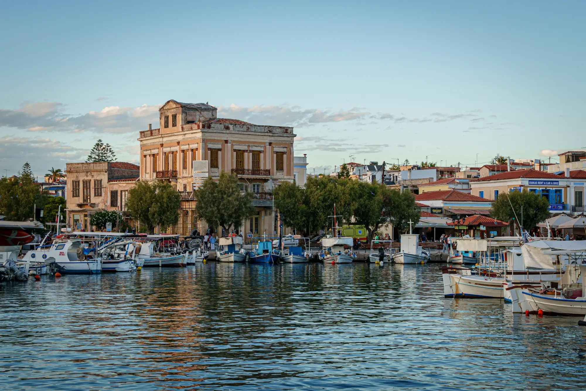 Small boats docked in the port of Aegina, Greece, with neoclassical buildings and trees along the waterfront.
