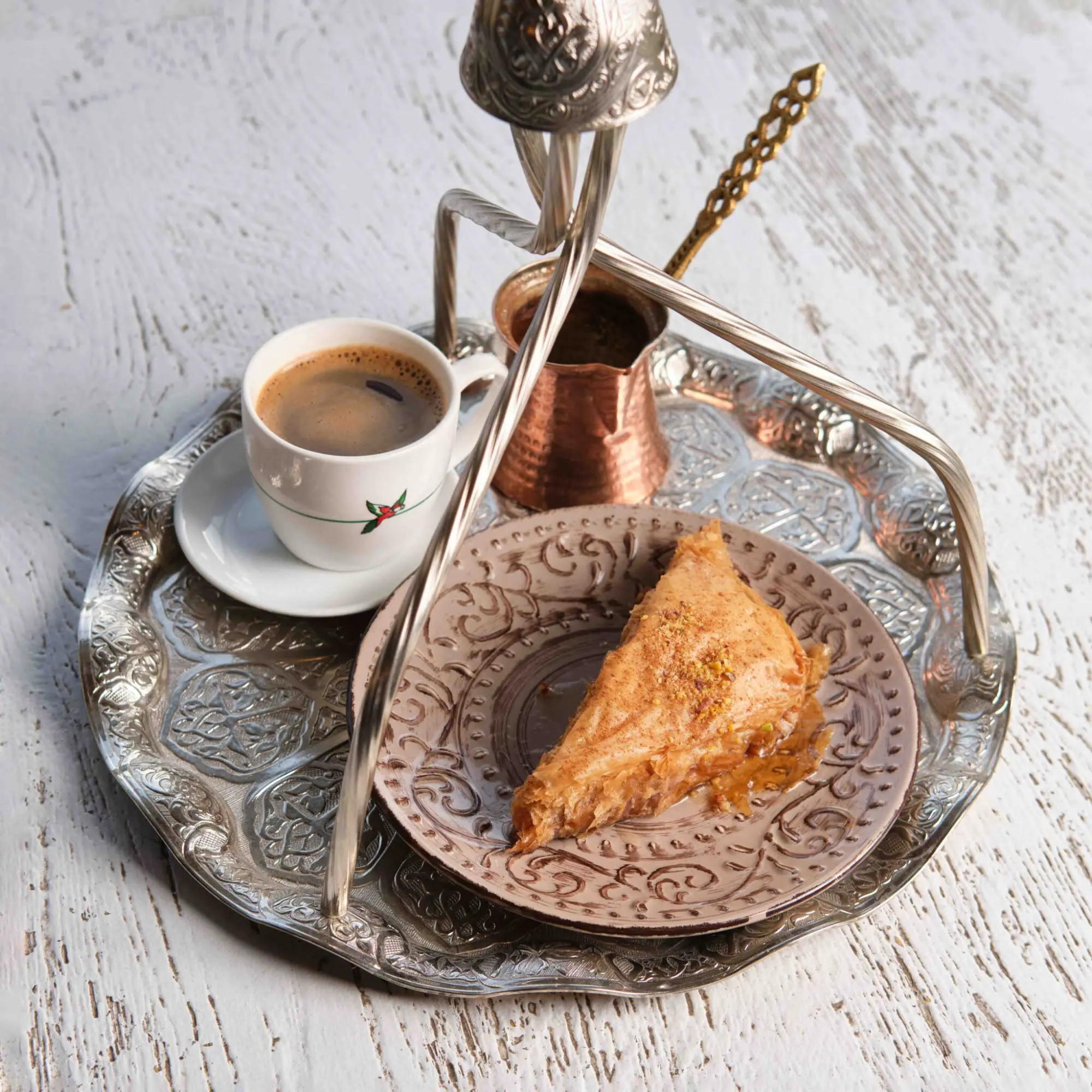 Hanging metal tray with a cup of Greek coffee, a copper pot, and a piece of baklava on a rustic white table.