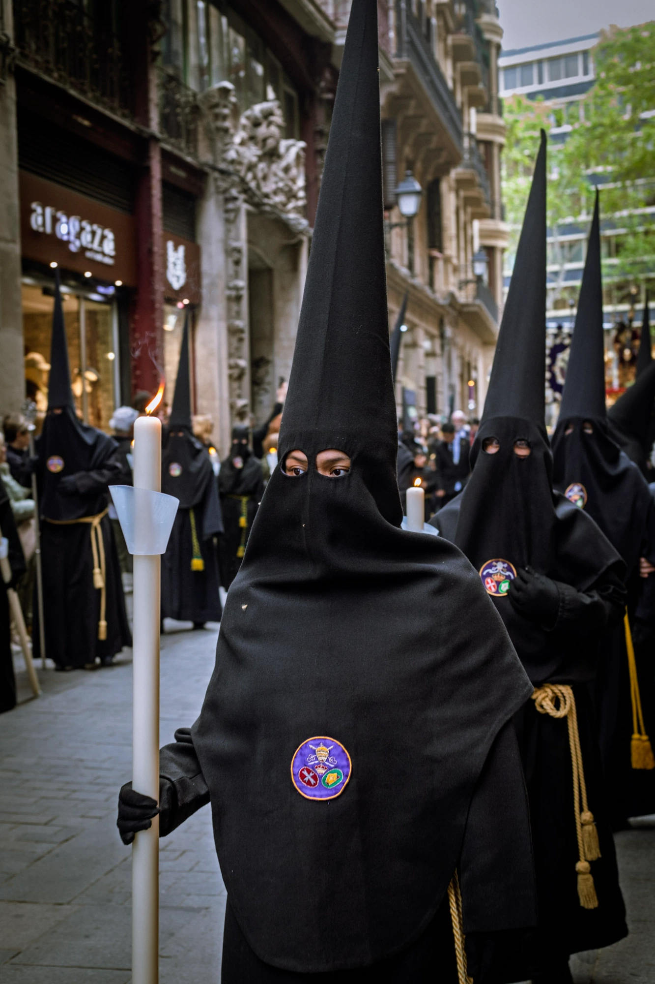 Person in black robe and conical hat holding a candle during a Holy Week procession in Barcelona.