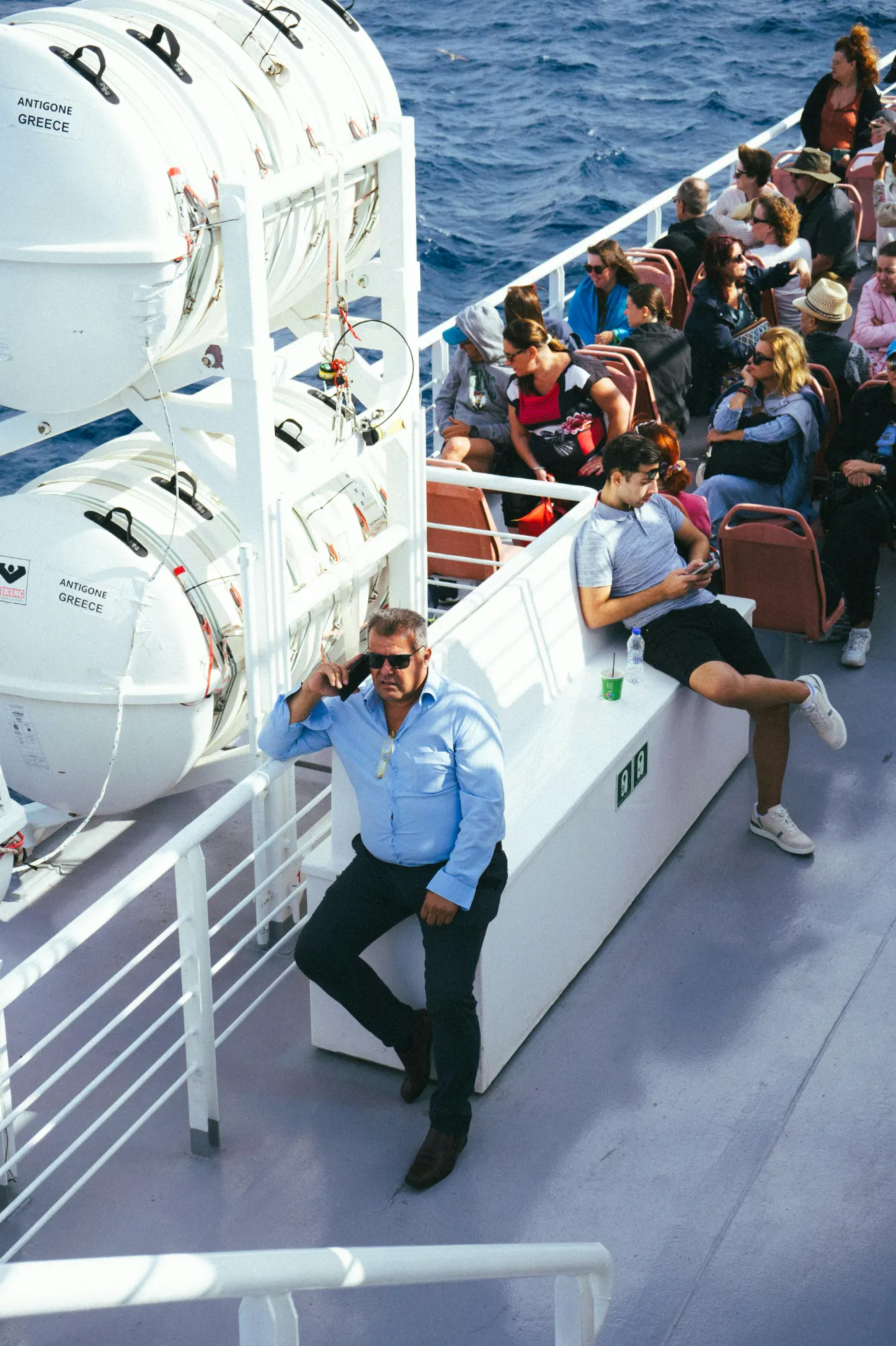Passengers relaxing on the sunny open deck of a ferry in Greece, with white life rafts visible.