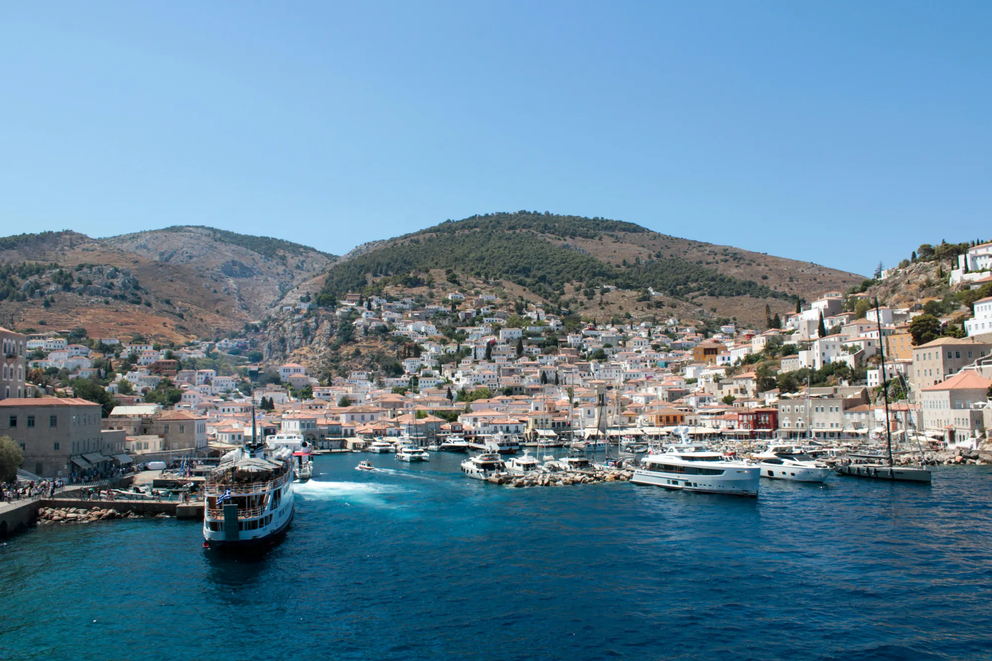 View of Hydra port in Greece with boats in the blue water and white houses built on the surrounding hills.