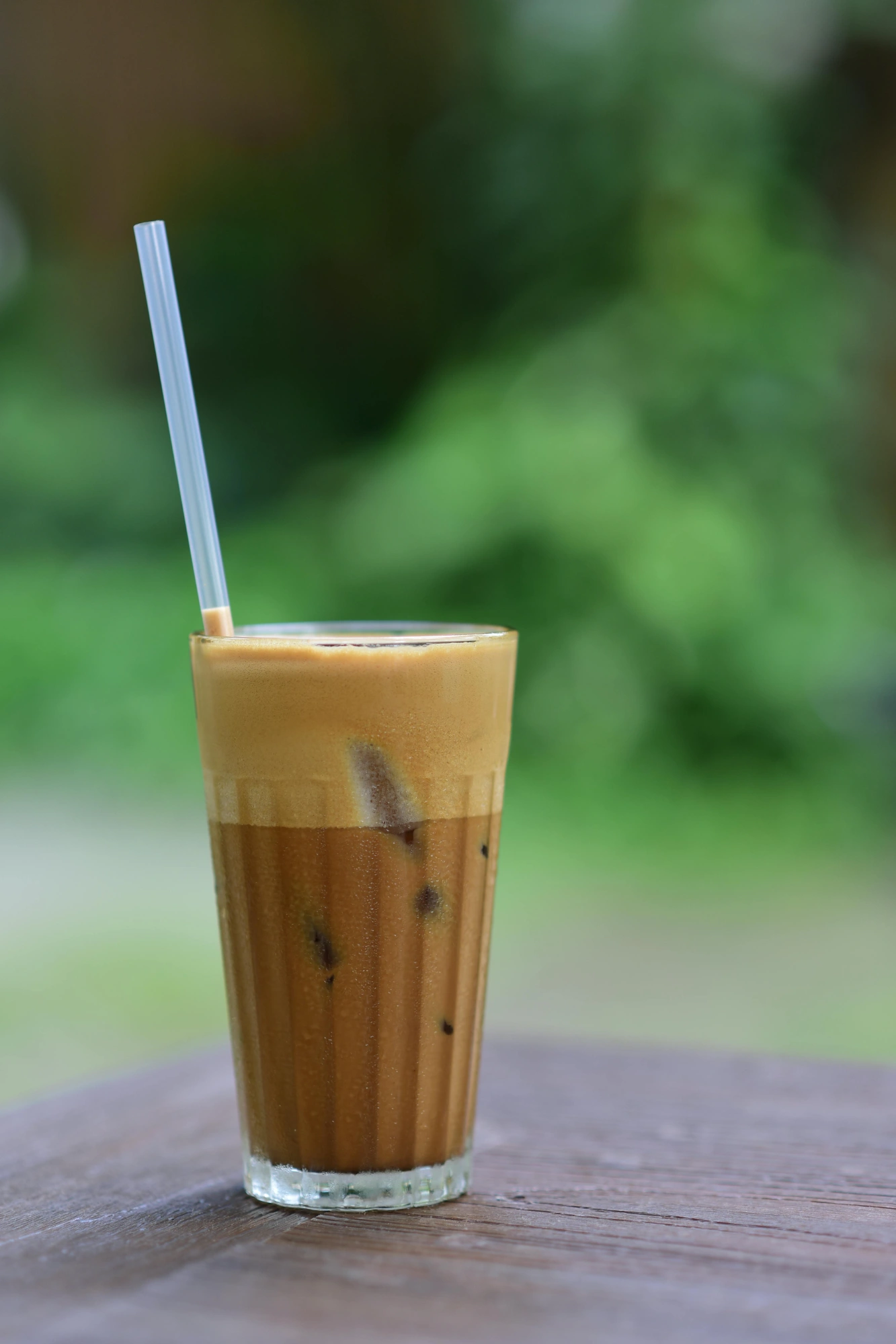 Tall glass of iced coffee with thick foam and a straw on a wooden table with a blurred green background.