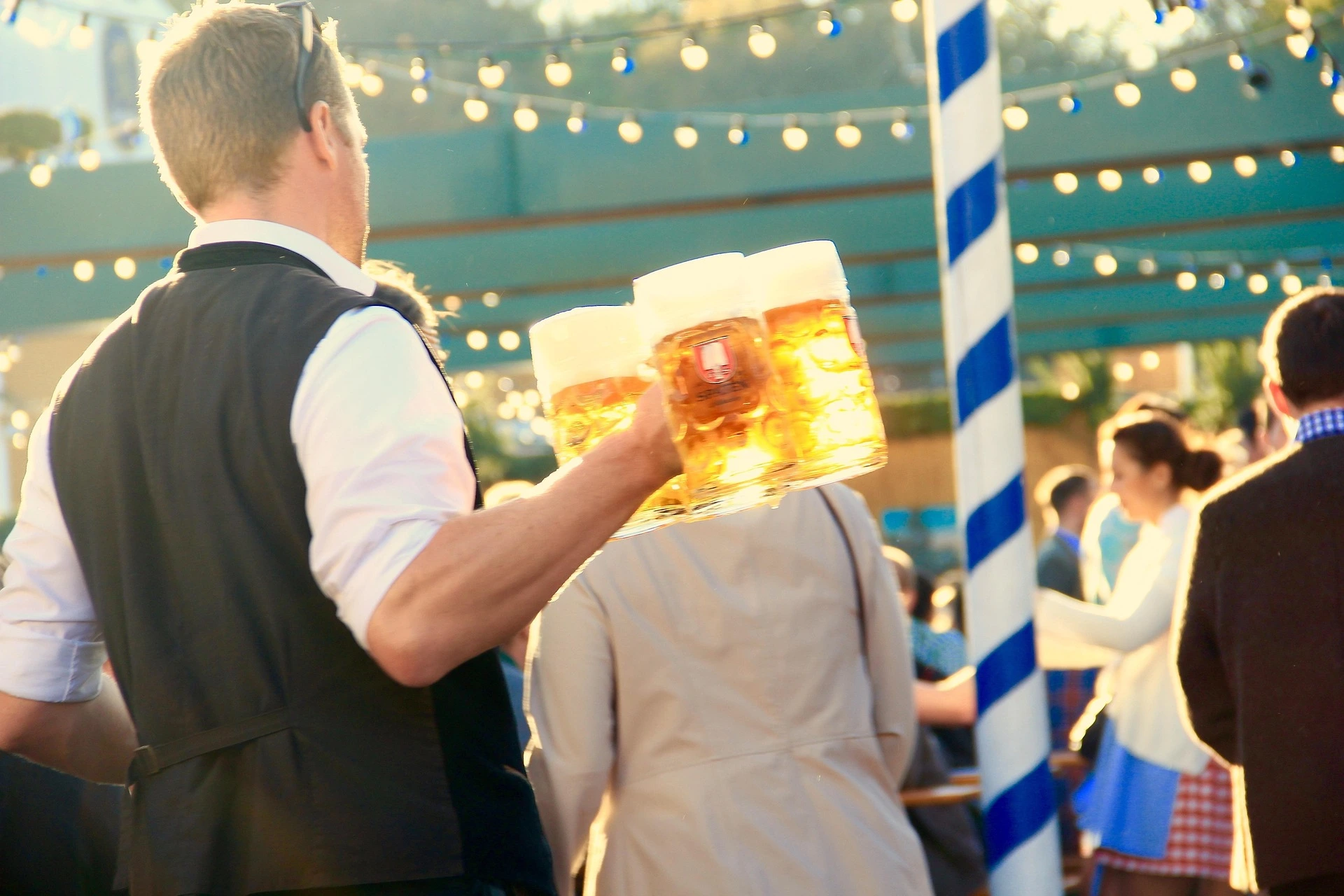 Ein Kellner trägt drei große Glaskrüge mit Bier auf einem Festgelände mit blau-weißem Mast.