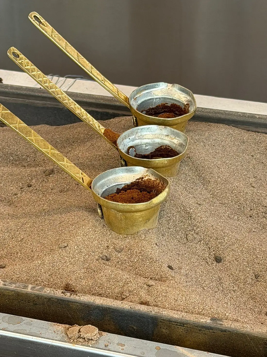 Three traditional long-handled brass pots containing coffee grounds, resting in heated sand.