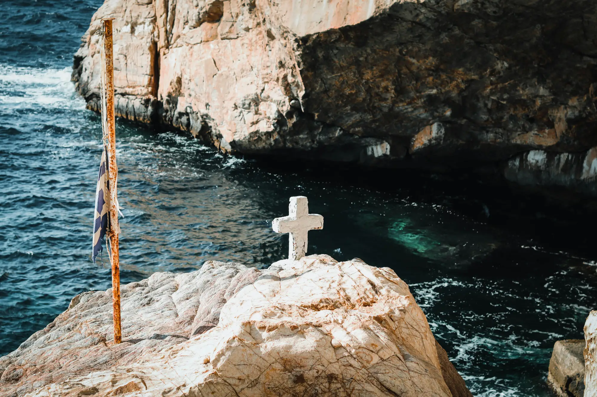 A white stone cross and a weathered Greek flag on a rocky cliff overlooking the sea in Syros.