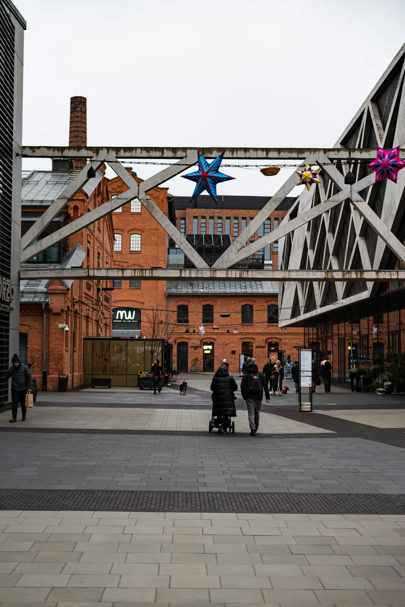 People walking in Koneser Center plaza in Warsaw under industrial beams with hanging star decorations.