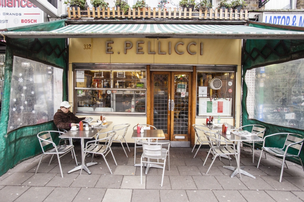 Fasada kawiarni E Pellicci w Londynie. Mężczyzna czyta gazetę przy stoliku na zewnątrz, pod zieloną markizą.