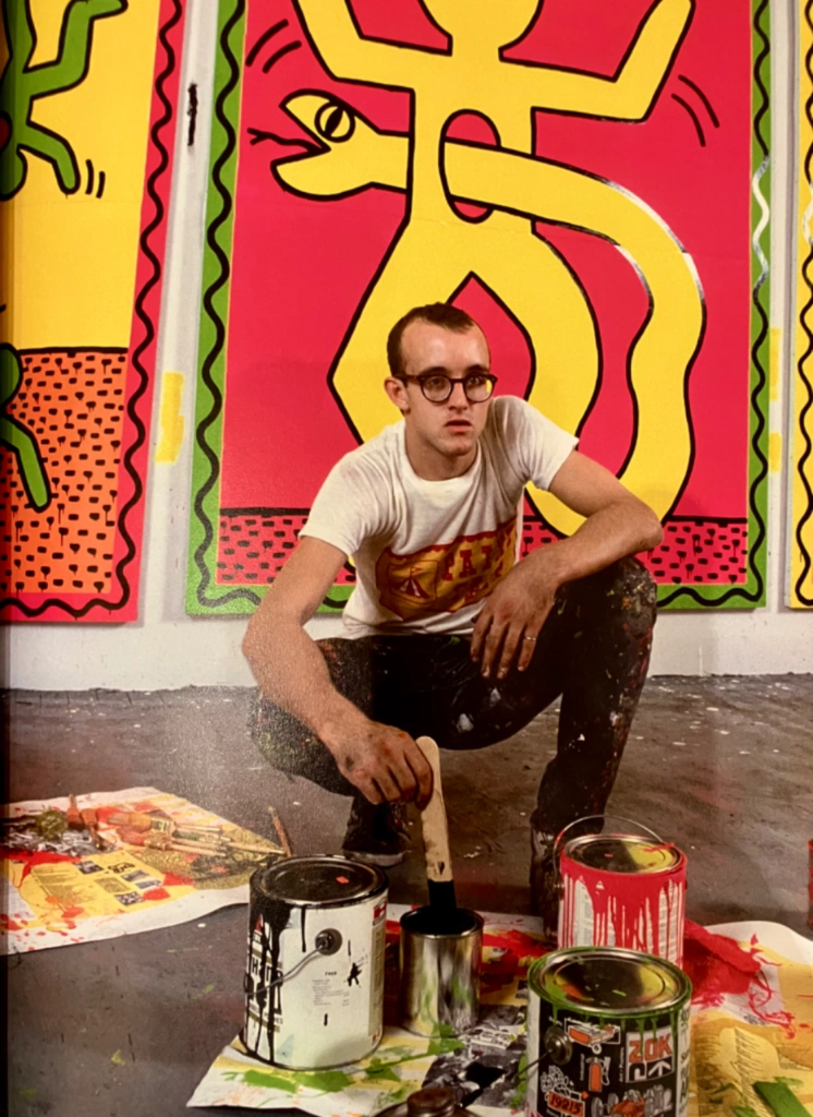 Artist Keith Haring squatting with paint cans in front of colorful pop art canvases in a studio.