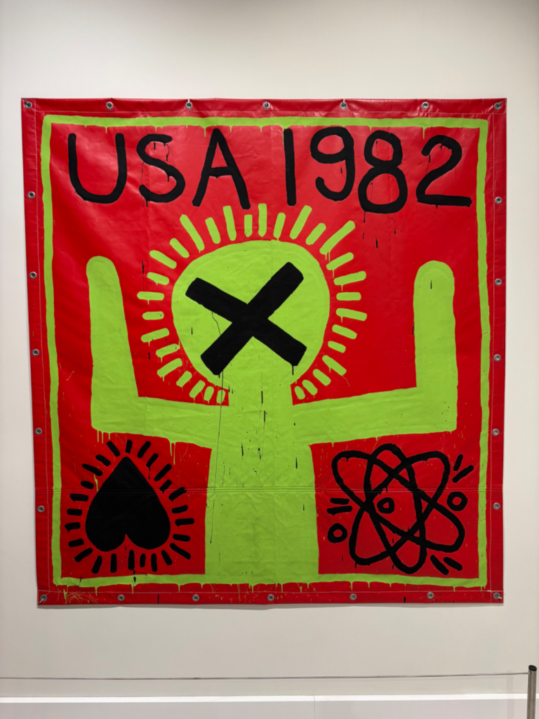 Red and green artwork by Keith Haring featuring a figure with an X face, a heart, an atom, and text USA 1982.
