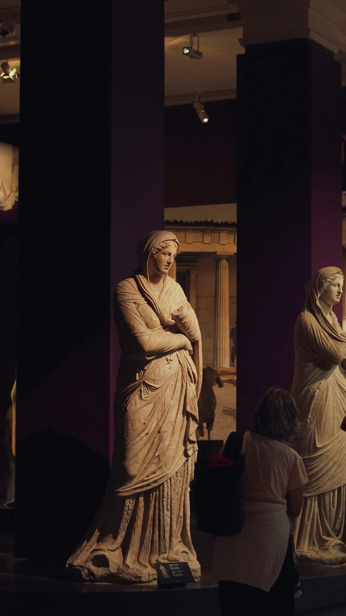 An ancient Greek marble statue of a woman in draped robes, viewed by a visitor in a softly lit museum gallery.