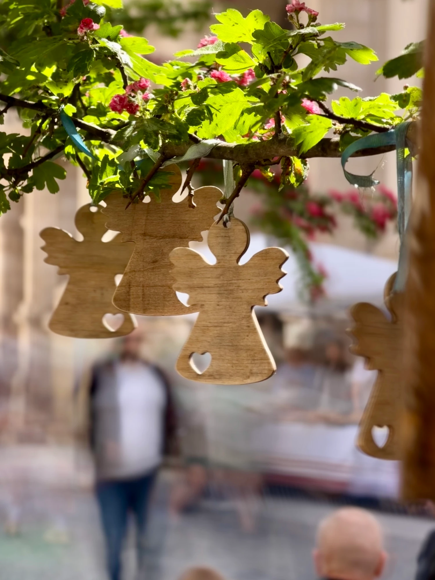 Wooden angel-shaped ornaments with heart cutouts hang from a blooming tree branch. The background is a blurry street scene.
