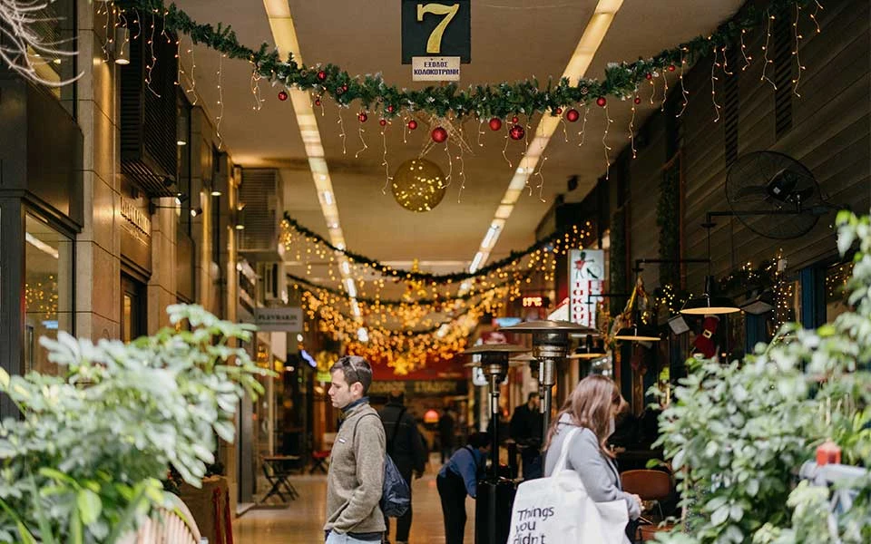 Pedestrians walking through an Athens shopping arcade decorated with holiday garlands and string lights.