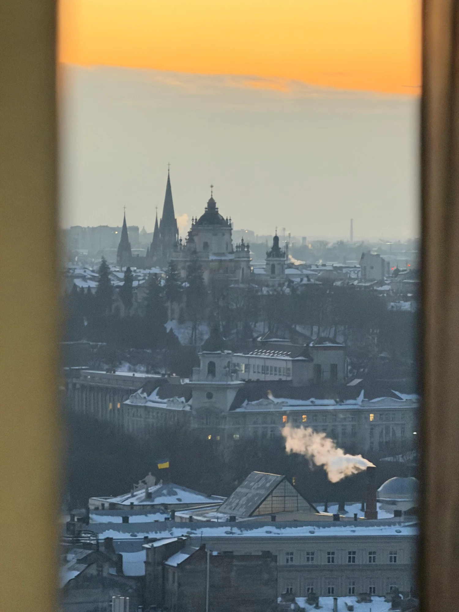 Lviv's skyline in winter at sunset, seen through a window, with snow-covered roofs and church spires in the distance.